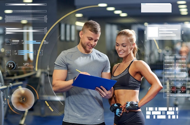 fitness, sport, exercising and people concept - smiling young woman and personal trainer with clipboard writing exercise plan in gym over virtual charts
