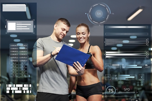 fitness, sport, exercising and people concept - smiling young woman and personal trainer with clipboard writing exercise plan in gym over virtual charts