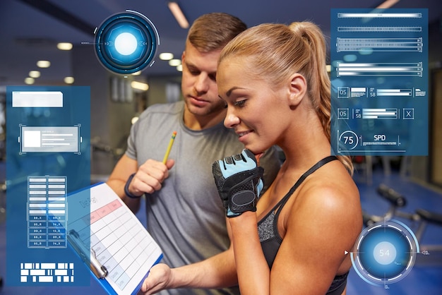 Photo fitness, sport, exercising and people concept - smiling young woman with personal trainer and exercise plan on clipboard in gym over virtual charts