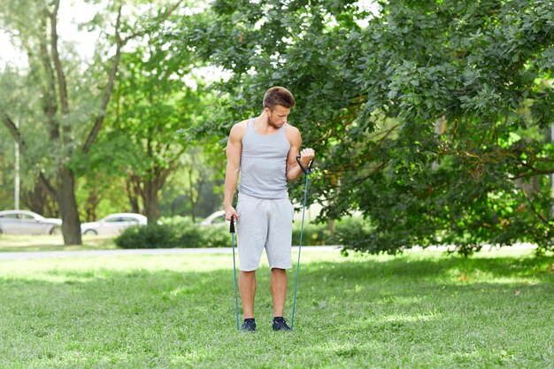 フィットネス、スポーツ、エクササイズ、トレーニング、ライフスタイルのコンセプト – 夏の公園でエキスパンダーを使って運動する若い男性