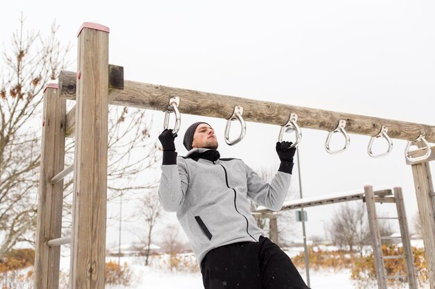 fitness, sport, exercising, training and people concept - young man doing pull ups on horizontal bar outdoors in winter