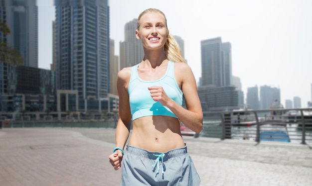 fitness, sport, friendship and healthy lifestyle concept - smiling young woman running or jogging over dubai city street background