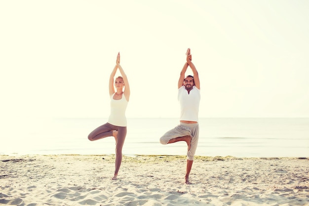 fitness, sport, friendship and lifestyle concept - couple making yoga exercises on beach