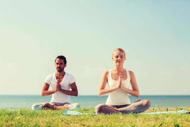 fitness, sport, friendship and lifestyle concept - smiling couple making yoga exercises sitting on mats outdoors