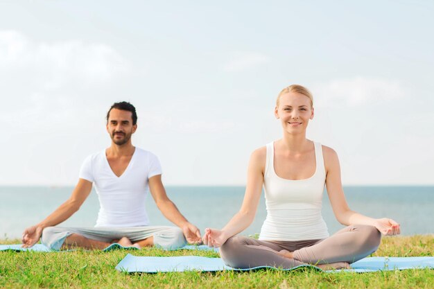 fitness, sport, friendship and lifestyle concept - smiling couple making yoga exercises sitting on mats outdoors