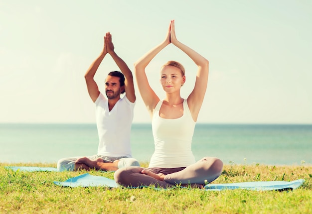 fitness, sport, friendship and lifestyle concept - smiling couple making yoga exercises sitting on mats outdoors