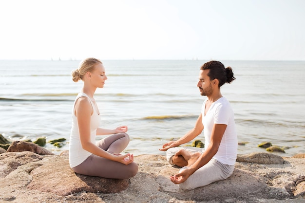 fitness, sport, friendship and lifestyle concept - smiling couple making yoga exercises sitting on sand outdoors