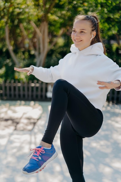 Fitness sport gezonde levensstijl concept Tevreden sportieve vrouw probeert op één been te balanceren houdt knie omhoog heeft intervaltraining of cardiotraining draagt witte sweatshirt leggings en sneakers