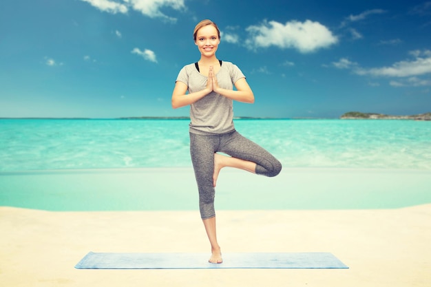 fitness, sport, mensen en een gezonde levensstijl concept - vrouw die yoga in boom maakt pose op mat over zee en lucht achtergrond