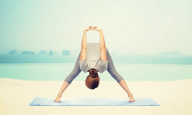 fitness, sport, mensen en een gezonde levensstijl concept - vrouw die yoga maakt in wijdbeens voorwaartse buiging pose op mat over infinity edge zwembad bij hotel resort achtergrond