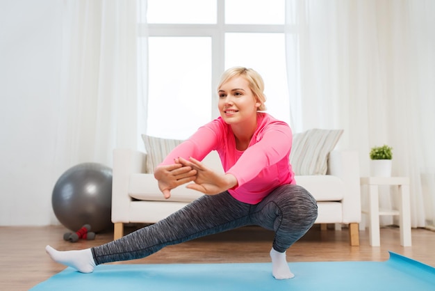 fitness, sport, people and healthy lifestyle concept - happy plus size woman stretching leg on mat at home