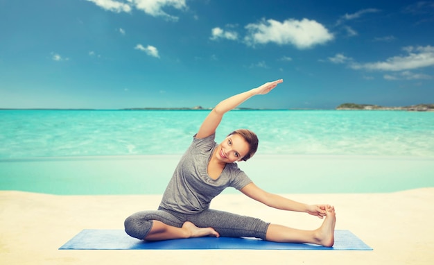 fitness, sport, people and healthy lifestyle concept - happy woman making yoga and stretching on mat over sea and sky background