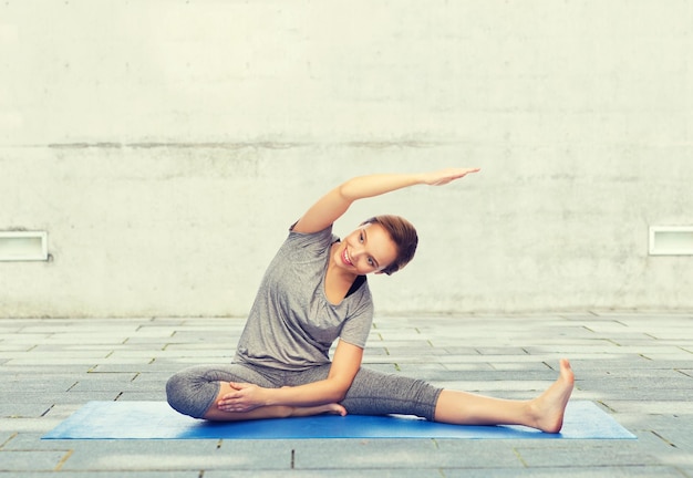 fitness, sport, people and healthy lifestyle concept - happy woman making yoga and stretching on mat over urban street background