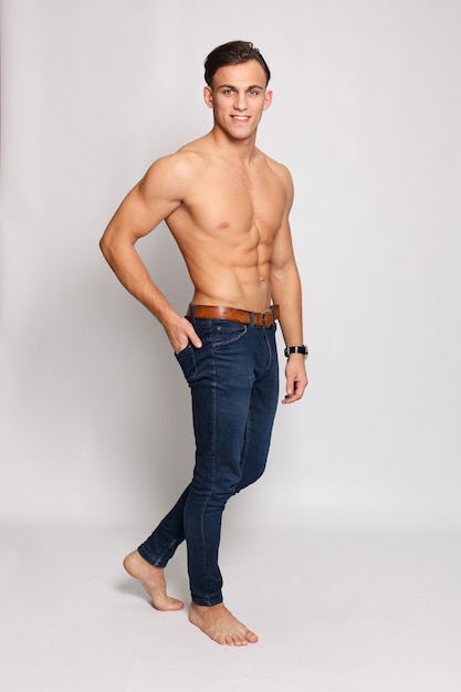 Fitness, sport, people, healthy lifestyle concept - Portrait of a young muscular man with naked torso standing with arms crossed against white wall. very muscular shirtless male model in jeans