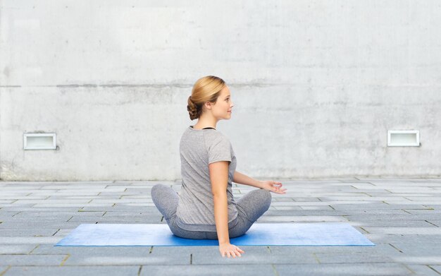 fitness, sport, people and healthy lifestyle concept - woman making yoga in twist pose on mat over urban street background