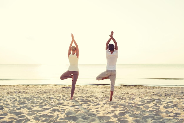 fitness, sport, people and lifestyle concept - couple making yoga exercises on sand outdoors from back