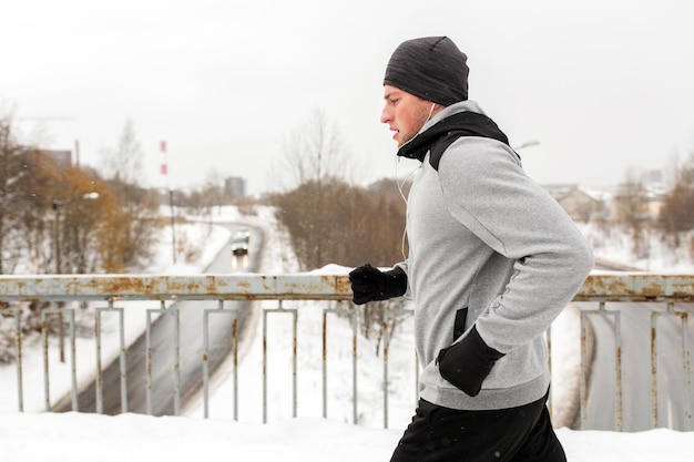 写真 フィットネス、スポーツ、人、音楽、健康的なライフスタイルのコンセプト – 雪に覆われた冬の橋に沿って走るイヤホンをした若者