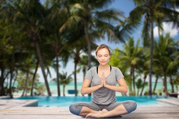 Photo fitness, sport, people, resort and healthy lifestyle concept - woman making yoga meditation in lotus pose over swimming pool and tropical beach with palm trees background