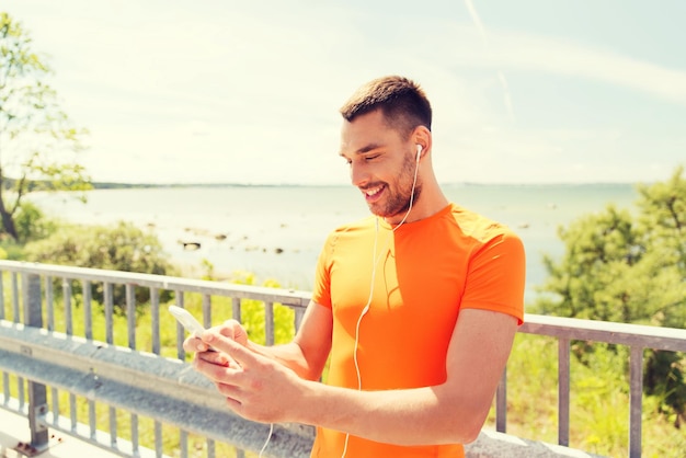 фитнес, спорт, люди, технологии и концепция здорового образа жизни - улыбающийся молодой человек со смартфоном и наушниками, слушающий музыку на летнем побережье