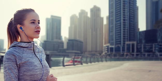 fitness, sport, people, technology and lifestyle concept - happy woman running and listening to music in earphones over dubai city street background