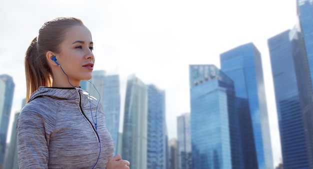 fitness, sport, people, technology and lifestyle concept - happy woman running and listening to music in earphones over singapore city skyscrapers background