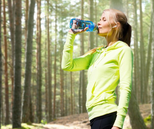 fitness, sport, people and thirst concept - happy woman drinking bottle water after doing sports over woods background