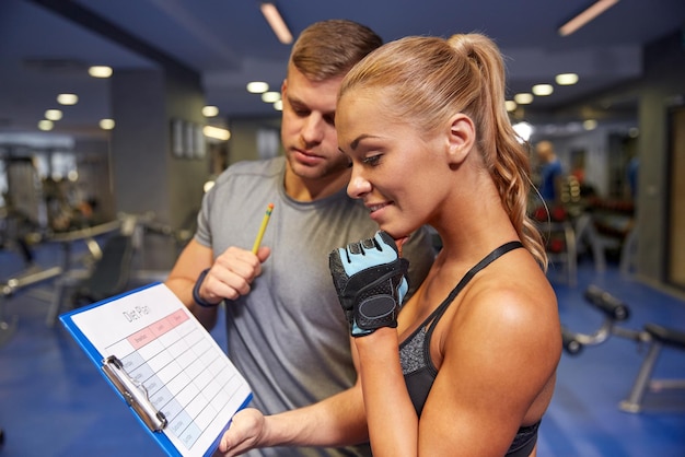 fitness, sport, sporten en dieetconcept - glimlachende jonge vrouw met persoonlijke trainer en oefeningsplan op klembord in gymnastiek