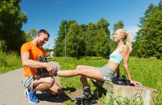 fitness, sport, summer, couple and healthy lifestyle concept - happy man helping woman to put on rollerblades outdoors