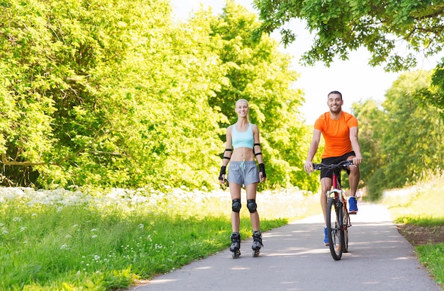 фитнес, спорт, лето, люди и концепция здорового образа жизни - счастливая пара с роликовыми коньками и ездой на велосипеде на открытом воздухе летом