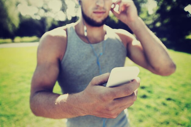 Photo fitness, sport, technology and lifestyle concept - close up of young man with smartphone and earphones listening to music at summer park