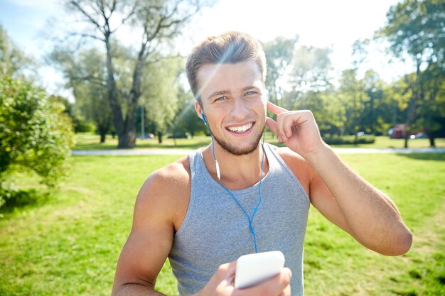 フィットネス、スポーツ、テクノロジー、ライフスタイルのコンセプト – 夏の公園でスマートフォンとイヤホンで音楽を聴く幸せな若い男性