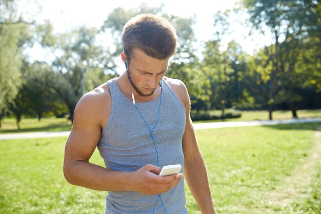 フィットネス、スポーツ、テクノロジー、ライフスタイルのコンセプト – スマートフォンと夏の公園で音楽を聴くイヤホンを持つ若者