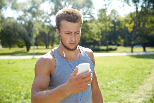 フィットネス、スポーツ、テクノロジー、ライフスタイルのコンセプト – スマートフォンと夏の公園で音楽を聴くイヤホンを持つ若者