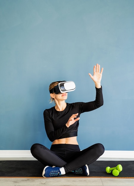 Fitness, sport and technology. Young athletic woman wearing virtual reality glasses sitting at fitness mat using VR interactive menu at blue background