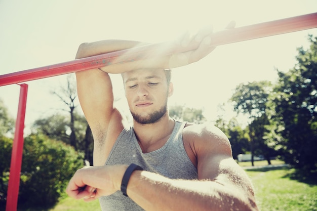 fitness, sport, training en lifestyle concept - gelukkige jonge man kijken naar hartslag horloge armband en oefenen op rekstok in zomer park