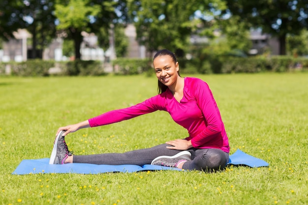 fitness, sport, training, park en lifestyle concept - glimlachende vrouw die het been op de mat buitenshuis uitrekt