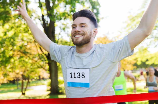 fitness, sport, victory, success and healthy lifestyle concept - happy man winning race and coming first to finish red ribbon over group of sportsmen running marathon with badge numbers outdoors