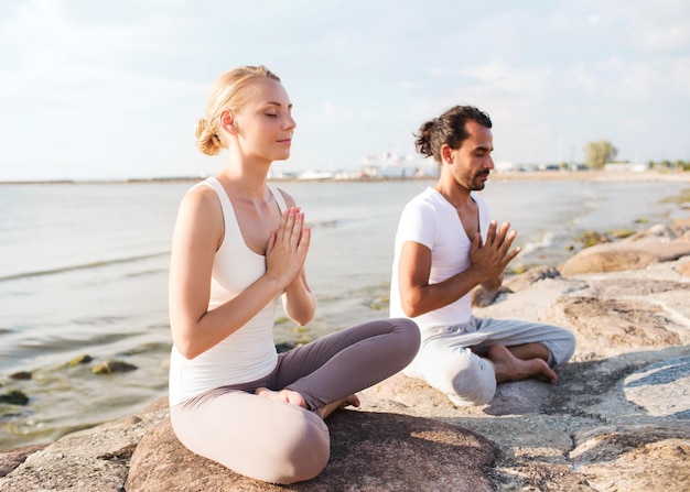 fitness, sport, vriendschap en lifestyle concept - glimlachend paar dat yoga-oefeningen maakt die buiten op zand zitten