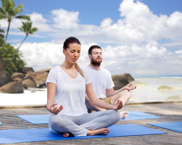 fitness, sport, yoga en een gezond levensstijlconcept - groep mensen die in lotushouding mediteren over exotische tropische strandachtergrond