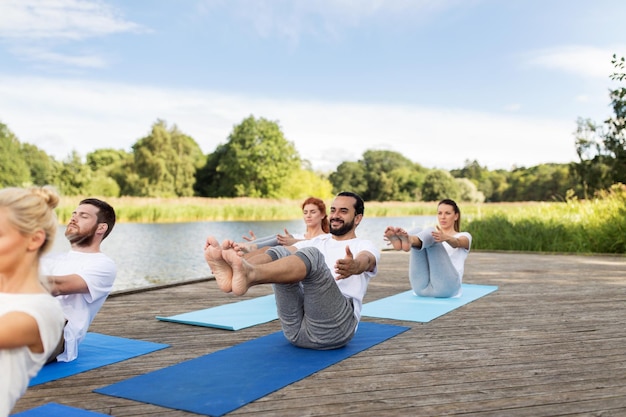 fitness, sport, yoga and healthy lifestyle concept - group of people making half-boat pose on mat outdoors on river or lake berth