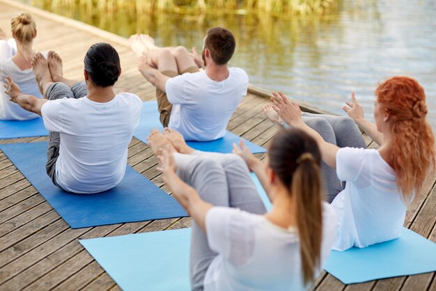 Fitness, sport, yoga and healthy lifestyle concept - group of people making half-boat pose on river or lake berth