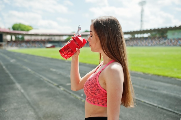 スタジアムの屋外スポーツでスポーツウェアのフィットネススポーティな女の子スポーツボトルのモックアップから水を飲む幸せなセクシーな女性