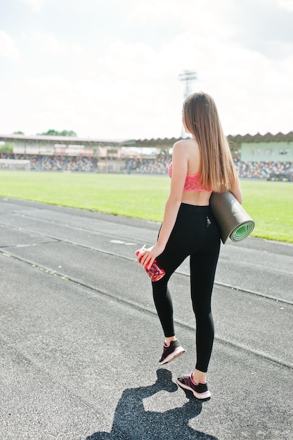 スタジアムの屋外スポーツでスポーツウェアのフィットネススポーティな女の子スポーツボトルのモックアップで幸せなセクシーな女性