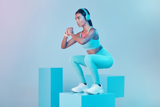 Fitness squat and woman with focus exercise and confident person against blue studio background Female model and athlete with headphones workout goal or balance for wellness or healthy lifestyle