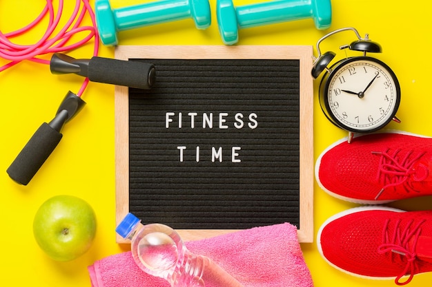 Photo fitness time and sport concept red sneakers apple jump rope dumbbells pink towel alarm clock and black letterboard with words fitness time on yellow background