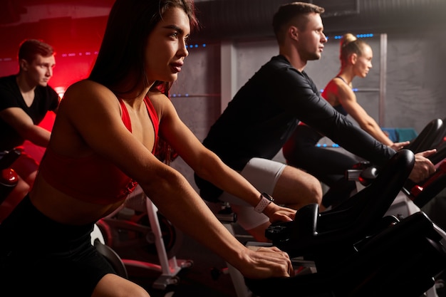 Fitness together on bicycles. young friends pedal on a stationary bike at the gym in red neon lighted space. cardio exercises on machine