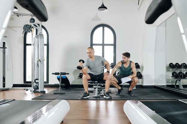 Fitness trainer asking client to keep legs wide open when doing biceps exercises with dumbbell