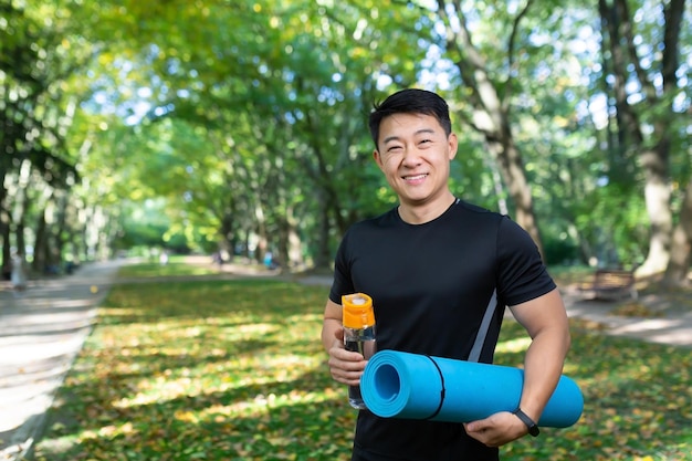 Fitness trainer looking at camera and smiling asian sports instructor with sports mat and bottle of