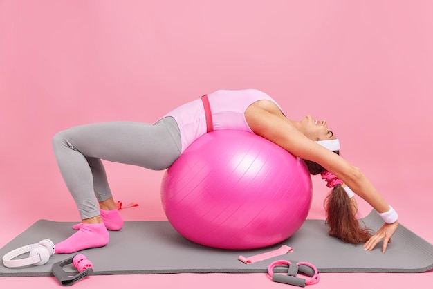 Fitness training concept. Slanke gezonde vrouw leunt op Zwitserse bal gekleed in sportkleding heeft gymnastiek op karemat doet buikspieroefeningen maakt gebruik van sportuitrusting geïsoleerd over roze achtergrond blijft gezond