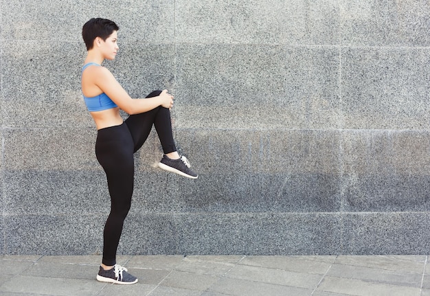 Fitness vrouw die haar benen strekt voordat ze naar buiten gaat. Jong slank meisje maakt aerobicsoefening, grijze muurachtergrond, kopieer ruimte